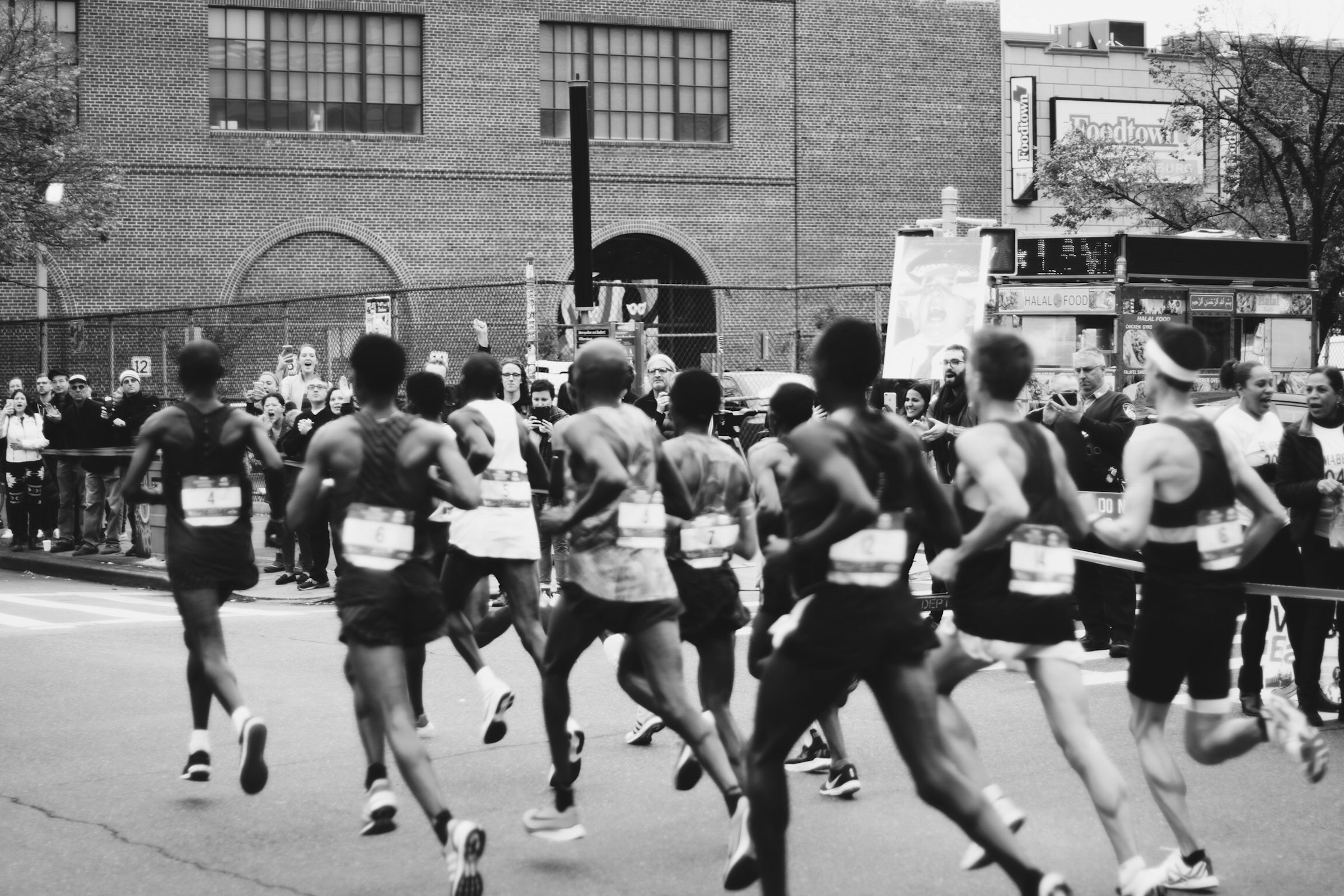 Maratonlöpare bakifrån i en klunga, publik och avspärrningar längs gatan, i svartvitt